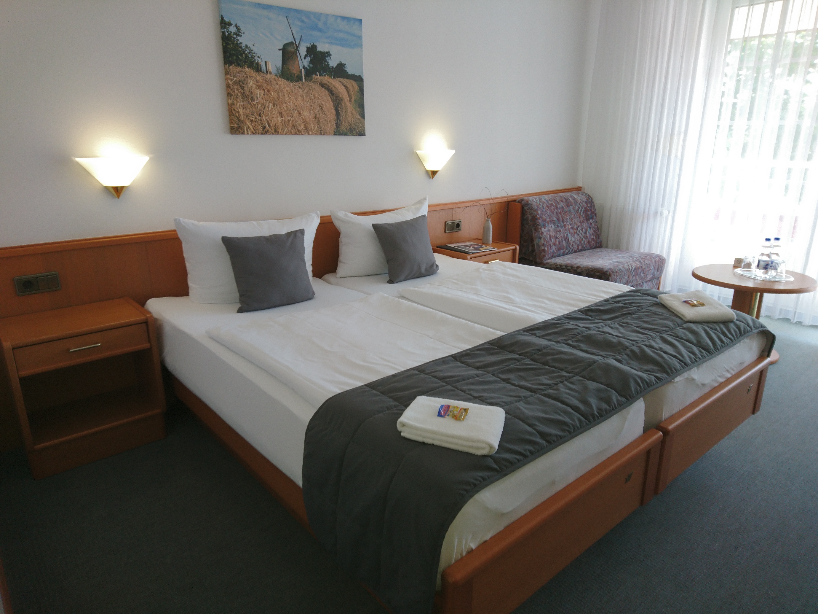 Hotelzimmer des Hotels Am Meilenstein mit Doppelbett, daneben Sitzgelegenheit und Beistelltisch, Blick durch große, bodentiefe Fenster nach draußen.